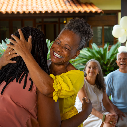 Mulher negra sorridente abraçando familiar em momento de carinho durante reunião de família ao ar livre.